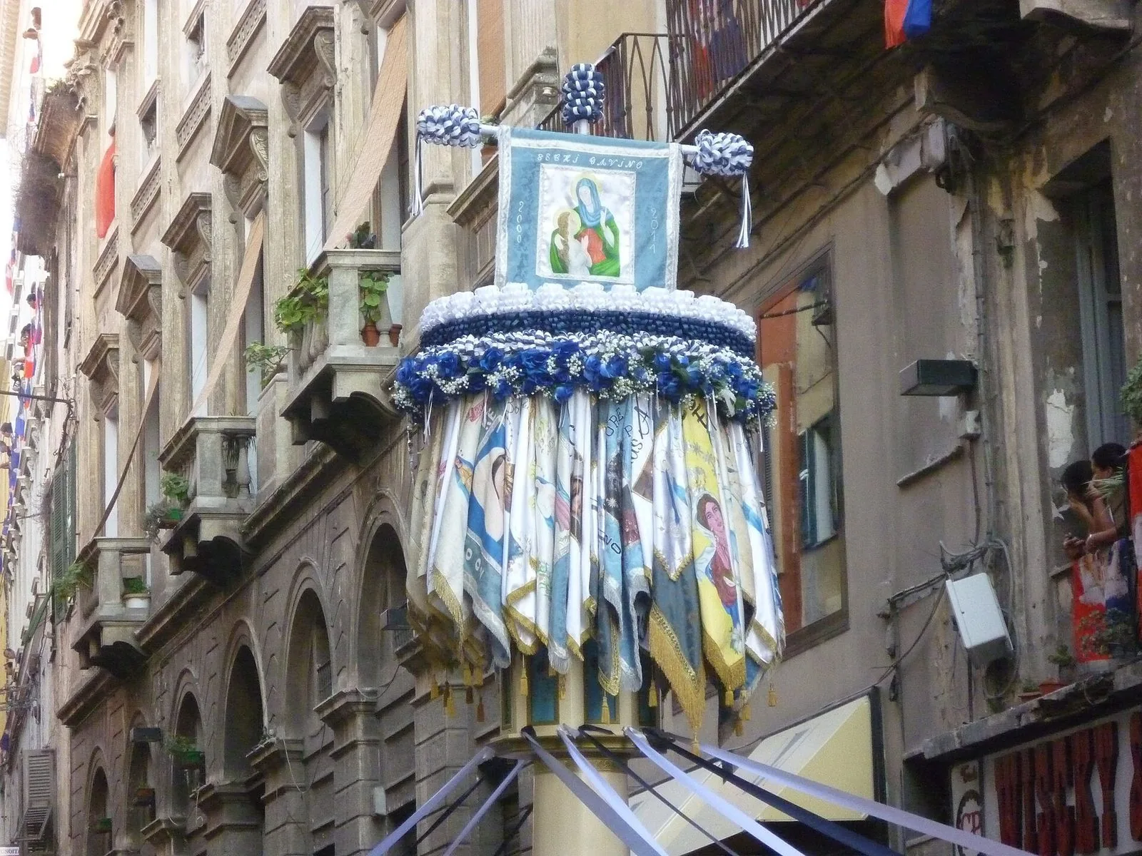 Sommità di un candeliere decorato con fiori blu, nastri e drappi con immagini sacre in una via del centro di Sassari
